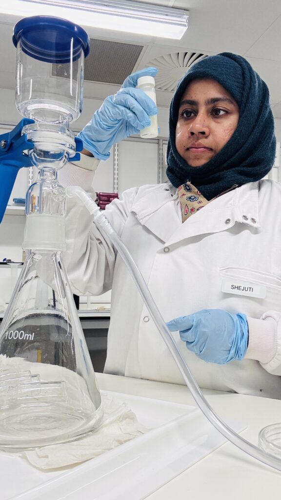 Student in lab coat examining a sample with lab apparatus in the foreground