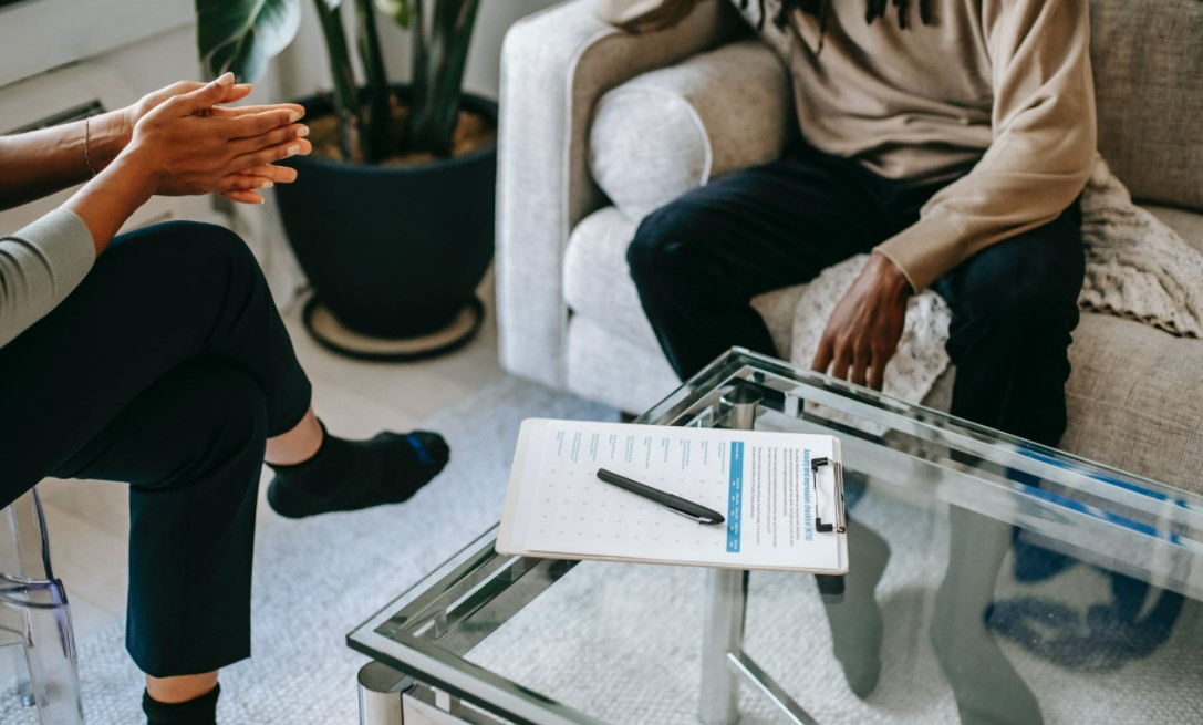 Two people sat on a sofa chatting, there is a clipboard on a glass table between them