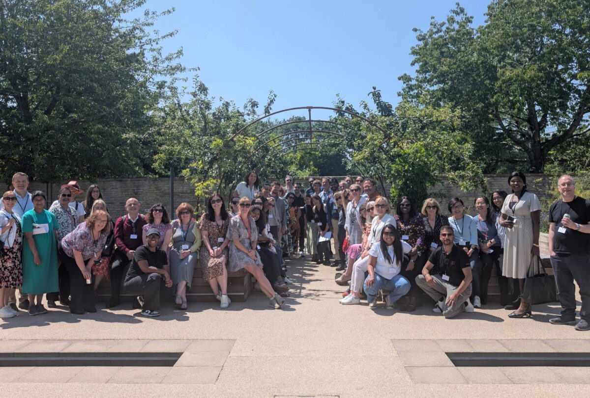 Large group of diverse professionals smiling and posing outdoors under a sunny sky, gathered around a garden archway at a work-related event or conference.