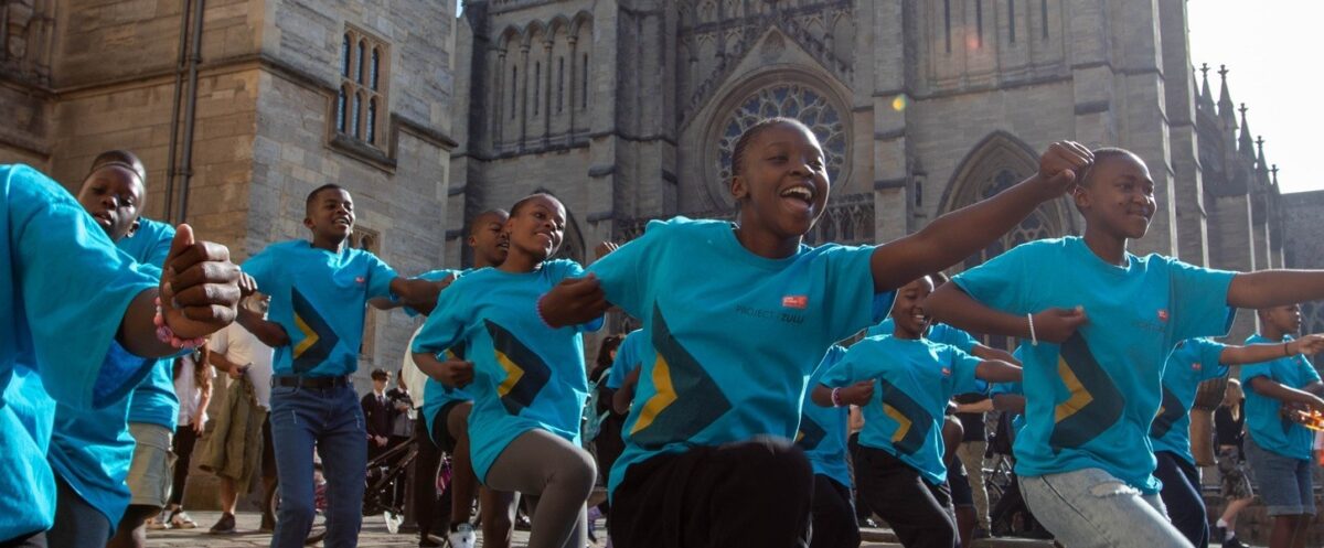 Children from the Project Zulu choir perform a traditional Zulu dance outside a cathedral, wearing bright blue tour T-shirts and smiling as they move in unison.