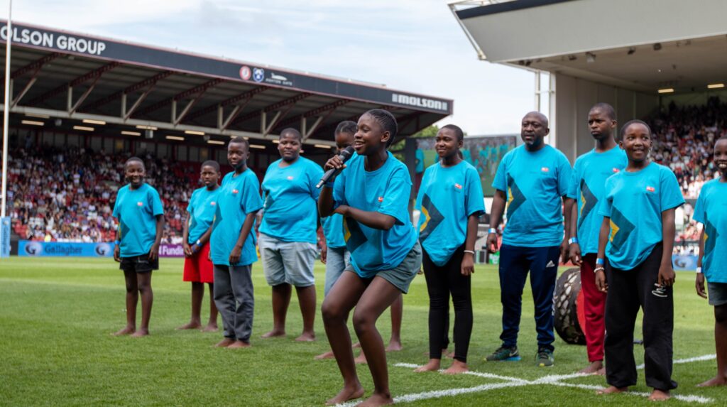 A young Project Zulu choir member sings into a microphone on the rugby pitch at Ashton Gate Stadium, with fellow choir members and teachers standing behind her in matching blue tour T-shirts.