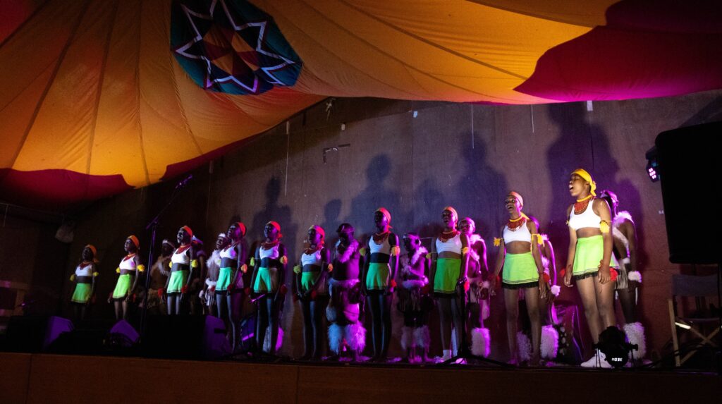 The Project Zulu choir performs on stage under a colourful marquee, dressed in traditional green, red, and white Zulu attire.