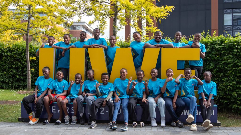 The 2025 Project Zulu choir poses for a group photo in front of the large yellow UWE Bristol sign, all wearing their blue tour T-shirts and smiling.