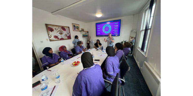 A group of Happy Care staff in purple uniforms sit around a table during a reablement training session, listening to a presentation with a slide displayed on a large screen.
