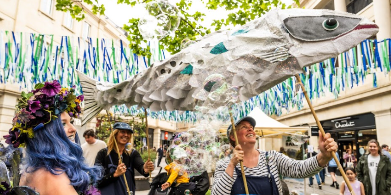 People frolicking in a metropolitan area. A beautiful two person fish puppet, long and thin with silver, sparkly scales and a woman dressed as nature with flowers and vines over her body and long hair occupy the foreground
