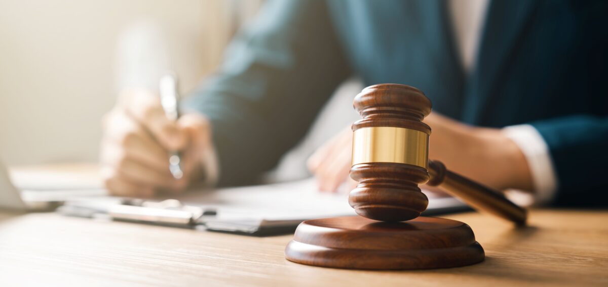 A picture of a wooden hammer used in law trials, there is a man writing in a notebook behind but it out of focus. the man is sat at a desk but you can only see his hands. the hammer is also on the desk in foreground.